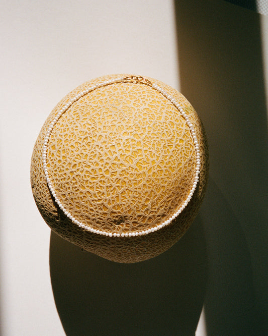 A close-up image of a delicate choker necklace with small Keshi beads and gold-filled components, displayed on a melon.