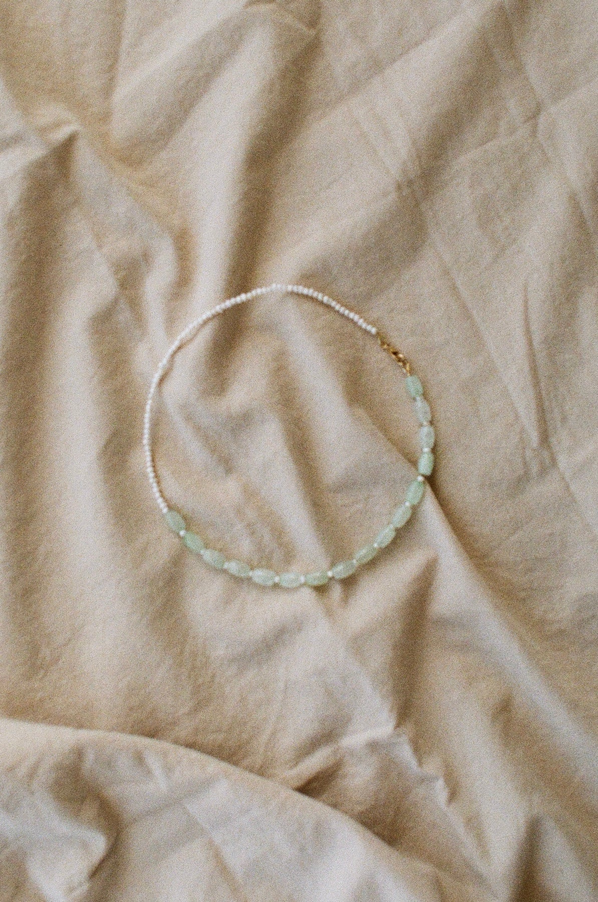 A choker necklace with Keshi pearls, greenish-blue oval-shaped jade beads and gold-filled components, displayed on a beige fabric surface.
