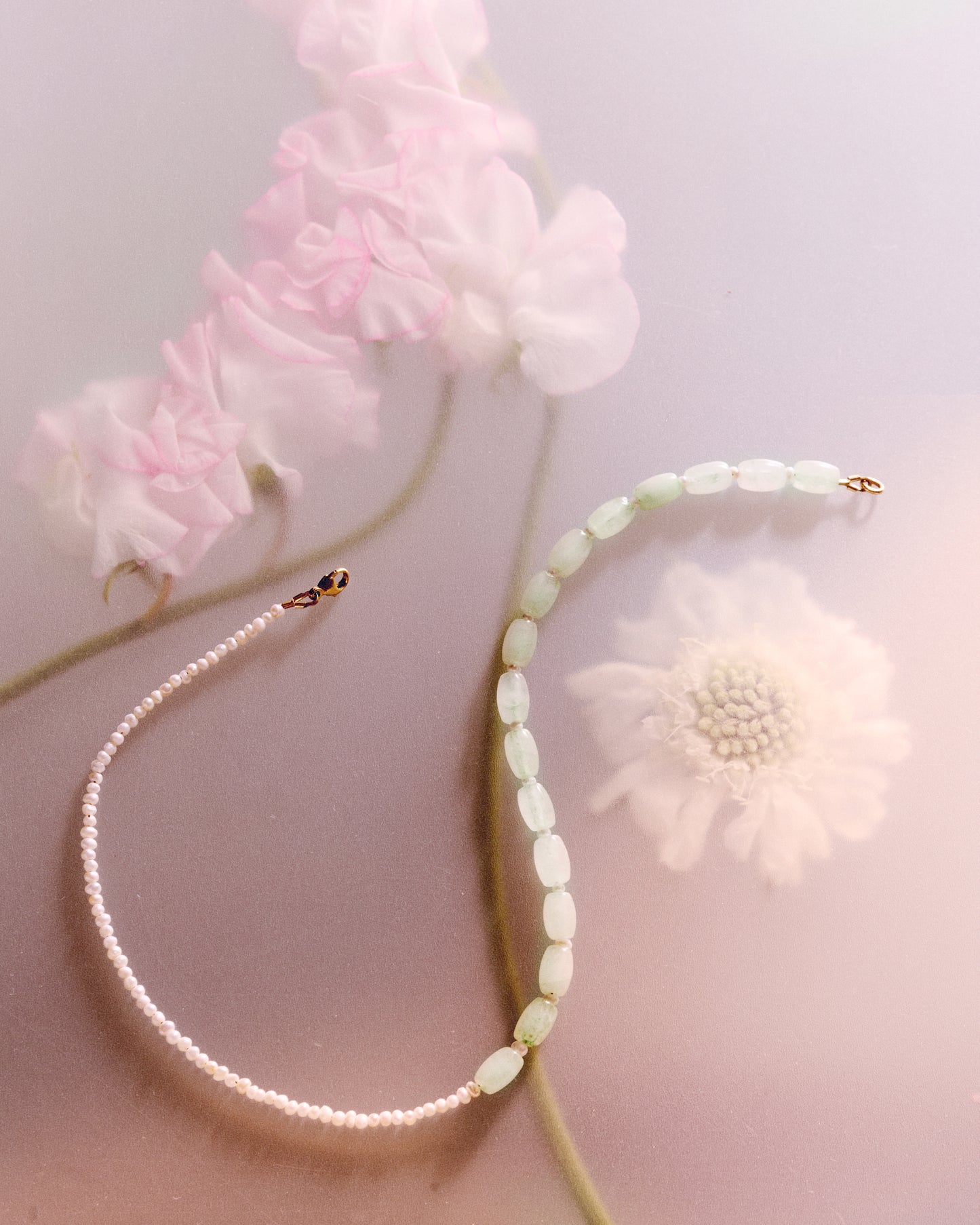 A necklace with half mini Keshi pearls and half minty blue, oval-shaped Jade beads displayed on a surface with flowers.